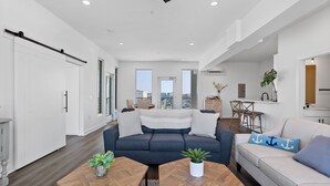 A modern living room with a sofa, coffee table, and a dining area. - Boardwalk Pent House | Ocean Views + Beach Access! (Carolina Beach)