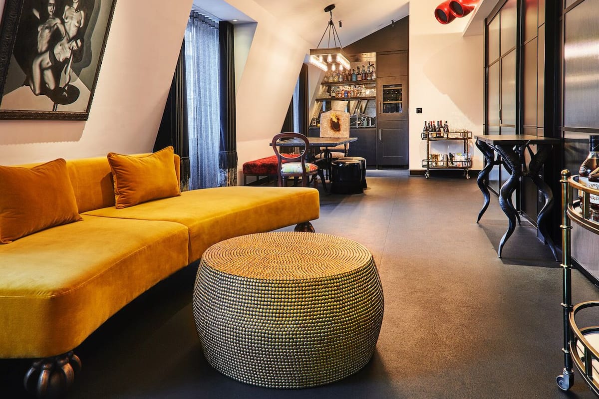 A modern living room with a yellow sofa, a round ottoman, and a bar area with shelves of bottles.