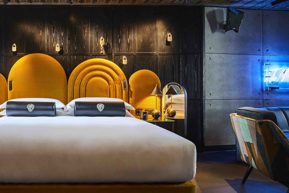 A hotel room with a large bed, multiple yellow chairs, a small table with a lamp, and a neon sign on the wall.
