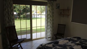 A room with a bed, two chairs, a sliding glass door to a garden, and a small shelf with items.