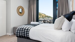 A modern bedroom with a large bed, a view of a cityscape, and a balcony with a chair. - Lush Sky Apartment With Ocean Views (Cape Town)