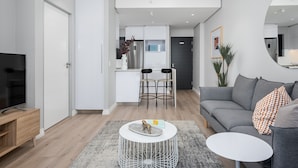 A modern living room with a grey sofa, a white coffee table, and a TV stand. A kitchen area is visible in the background with a fridge and bar stools. - Lush Sky Apartment With Ocean Views (Cape Town)