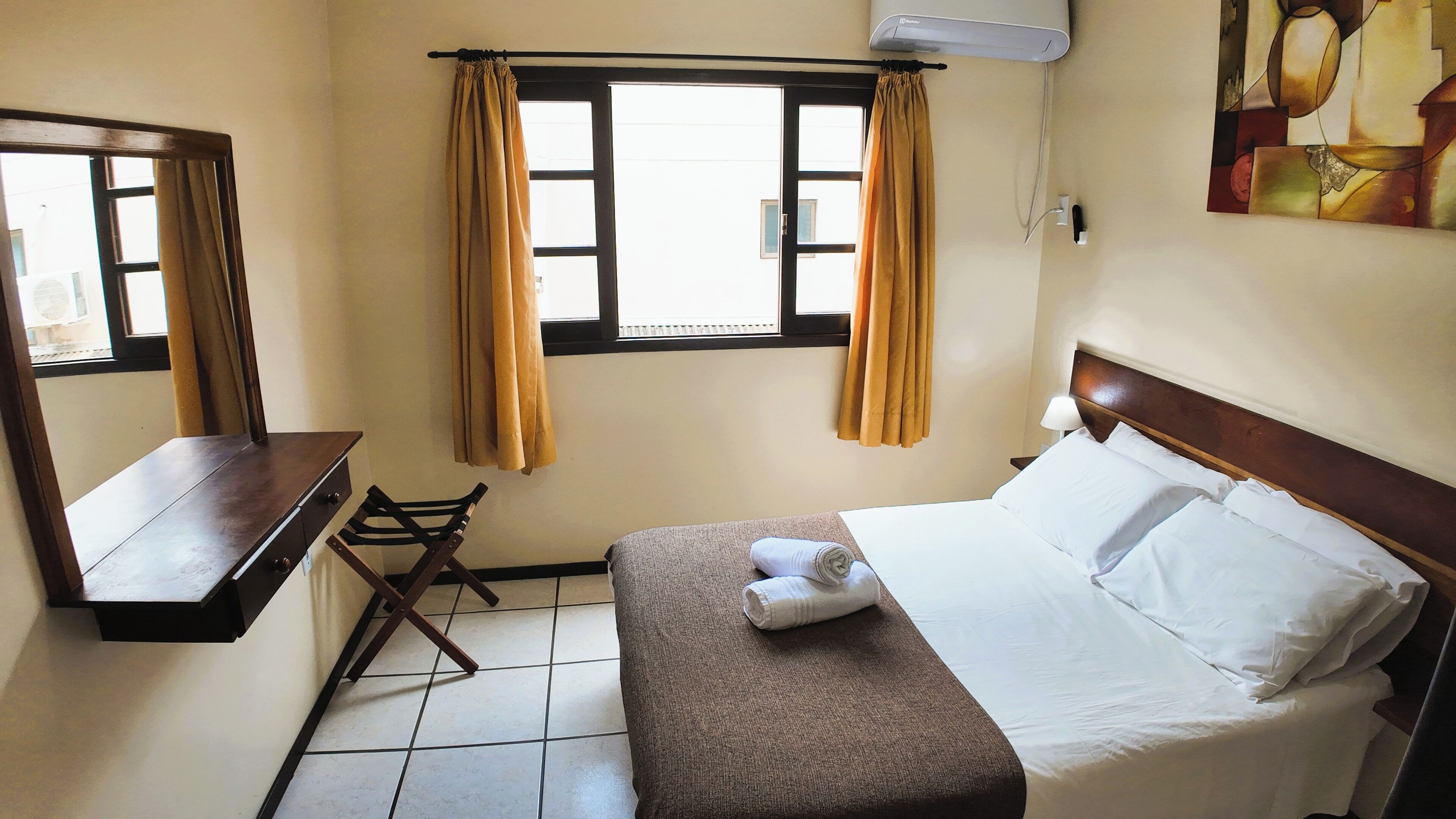 Une chambre d’hôtel avec un lit, un petit bureau, une chaise, une fenêtre avec des rideaux et un tableau au mur.