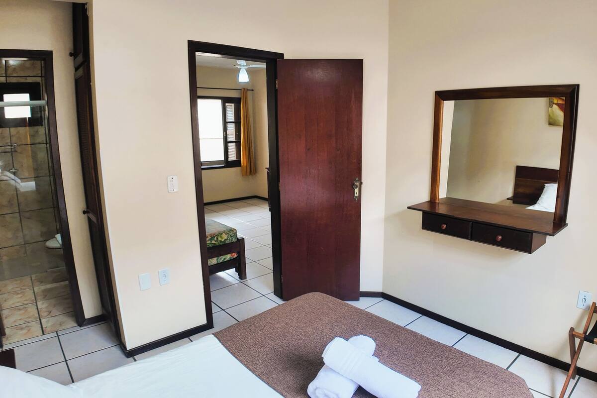 Une chambre d’hôtel avec un lit, une porte en bois, un miroir, une petite table et une salle de bain visible à travers une porte ouverte.