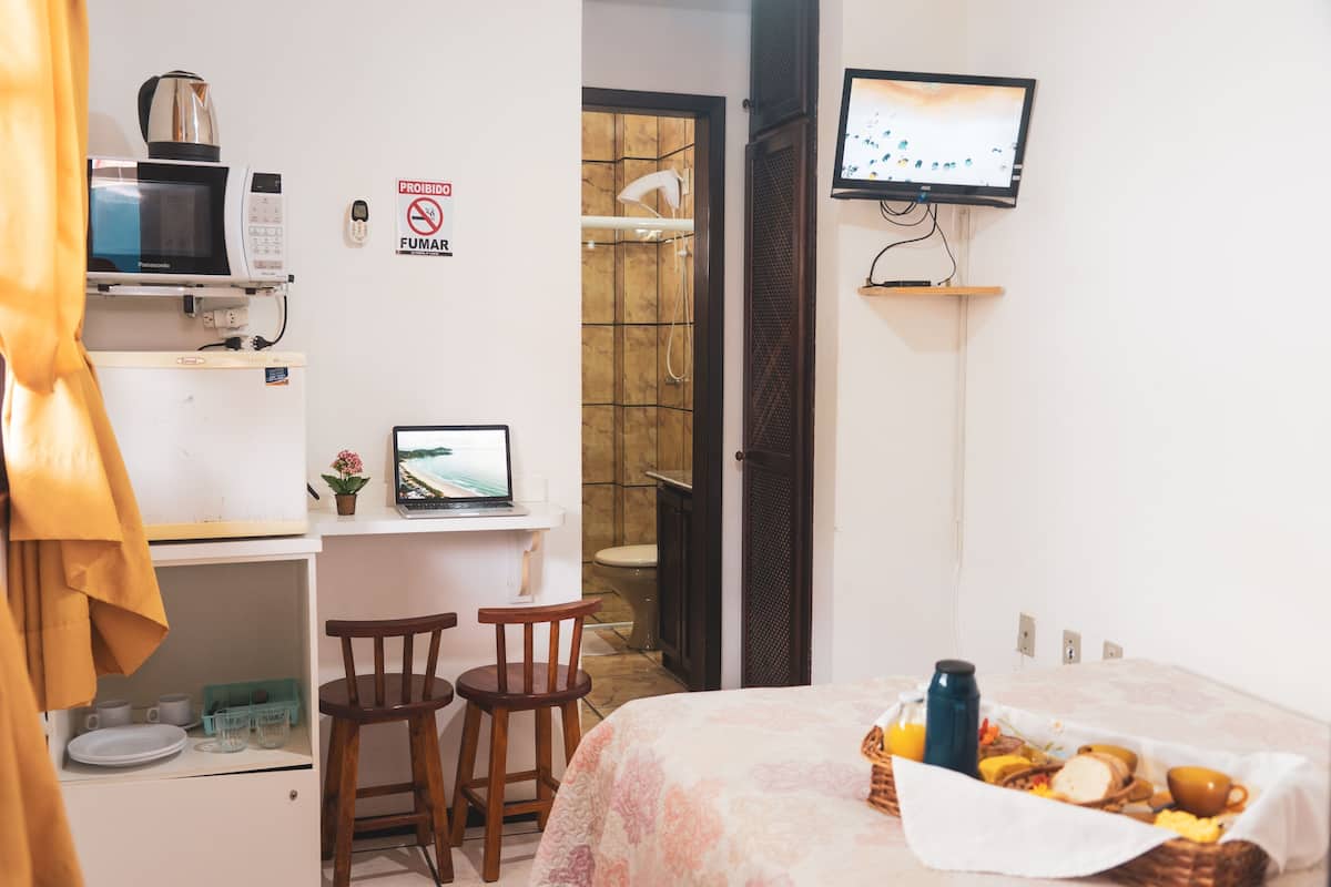 Une petite pièce comprenant un lit, un bureau avec un ordinateur, un four à micro-ondes et un téléviseur. On peut apercevoir un espace douche à travers une porte ouverte.