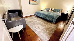A bedroom with a bed, a chair, a small table, a rug, and a painting on the wall. - Stay Roanoke | Ground-floor gem | walk to it all (Roanoke)