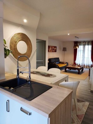 Una cocina moderna con encimera negra e integrada, una zona de comedor con mesa y sillas blancas, un salón con sofá y mesa de centro, y una gran decoración circular en la pared.