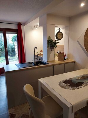 A compact kitchen with a sink, a window with red curtains, a clock on the wall, and a dining area with a table.