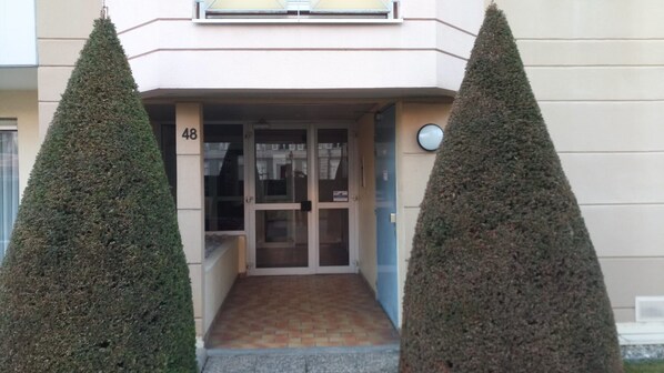 Entrada de un edificio con puerta de cristal, el número '48' en la pared y dos setos podados a cada lado.