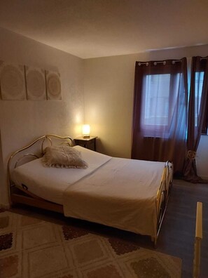 Una cama individual con cabecera, una mesita de noche con lámpara, una ventana con cortinas y una alfombra estampada.