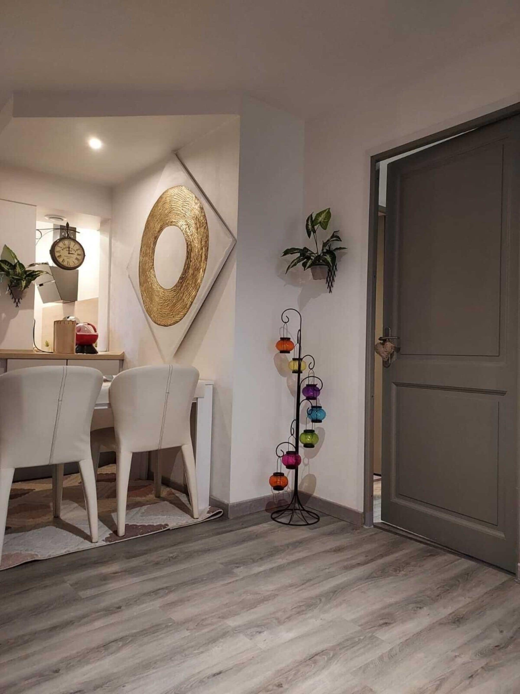 Un coin repas avec une table et des chaises blanches, une horloge murale, un miroir décoratif et une plante accrochée au mur.