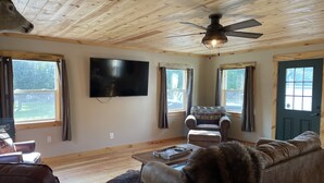 Un salon chaleureux avec un plafond en bois, un téléviseur fixé au mur, un canapé en cuir, un fauteuil à motifs et une table basse en bois.