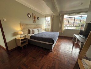 A bedroom with a wooden floor, a bed with a dark blanket, a nightstand with a lamp, a TV on a wooden stand, and a window with curtains. - Small loft for 4 people (Cusco)