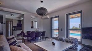En moderne stue med sofa, sofabord og spiseplads med stole. Der er et stort vindue med udsigt til en pool.