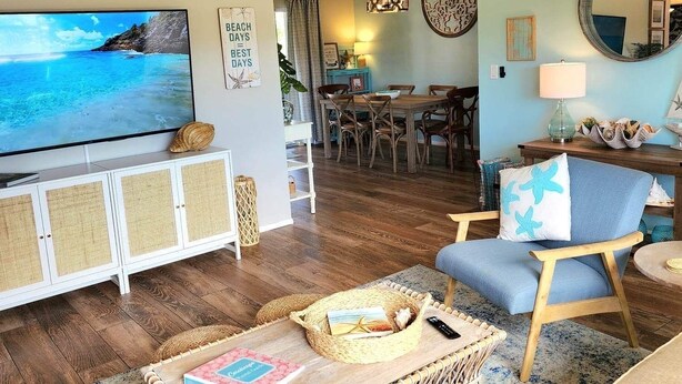 A living room with a television, wicker furniture, and a dining area in the background.