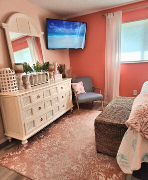 Ein Wohnzimmer mit einer weißen Kommode, einem an der Wand montierten Fernseher, einem grauen Sessel und einem gemusterten Teppich.