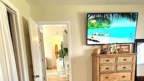 A hotel room with a wooden dresser, a flat-screen TV mounted on the wall, and a hallway leading to another room.