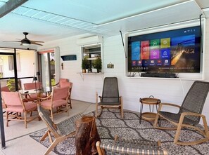 Une pièce dotée d’un grand téléviseur fixé au mur, d’un ventilateur de plafond et de plusieurs options d’assises, notamment des fauteuils en osier et une table.