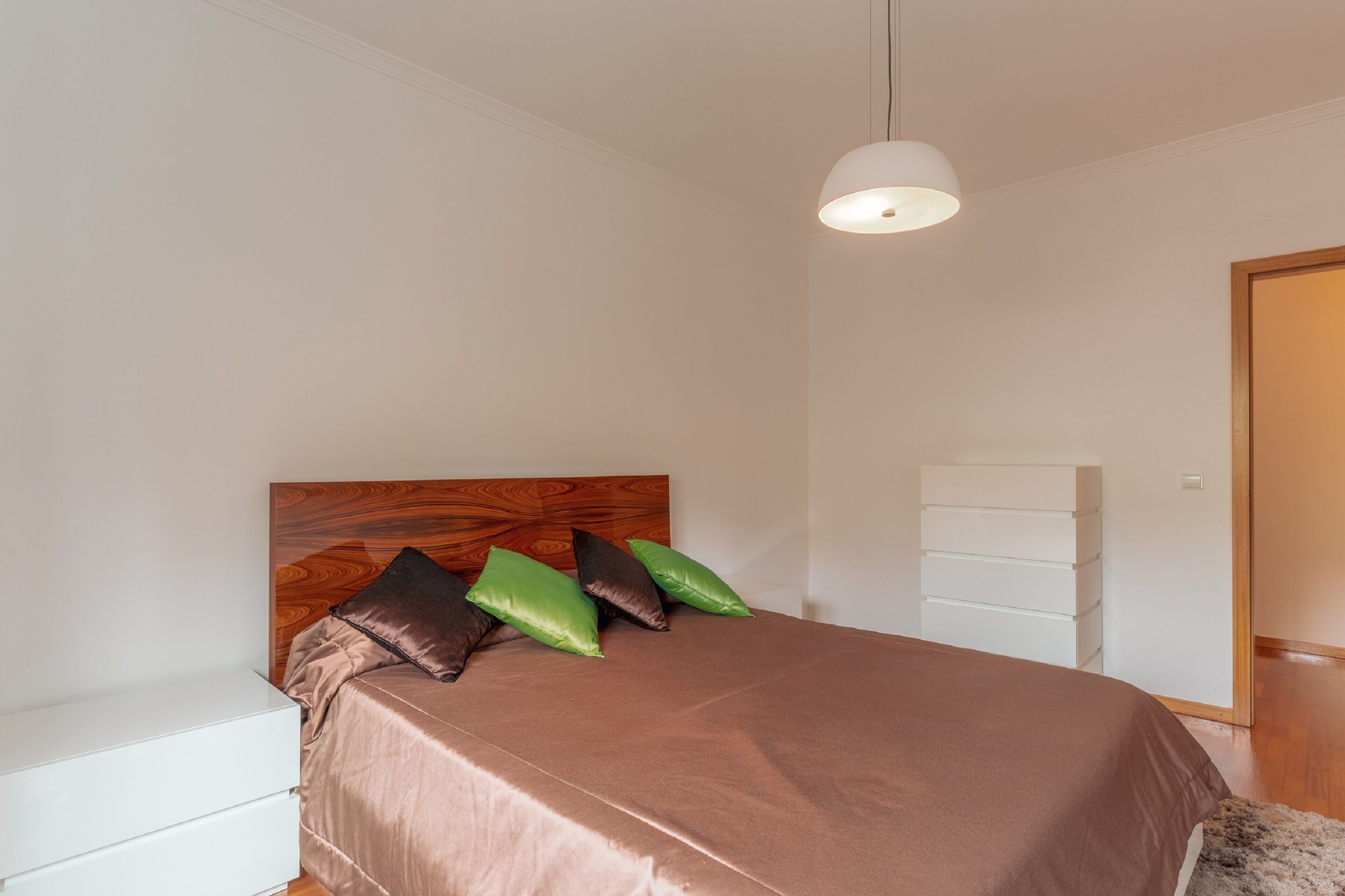 A bedroom with a wooden headboard, a bed with brown bedding, green and brown pillows, a white bedside table, and a white dresser.