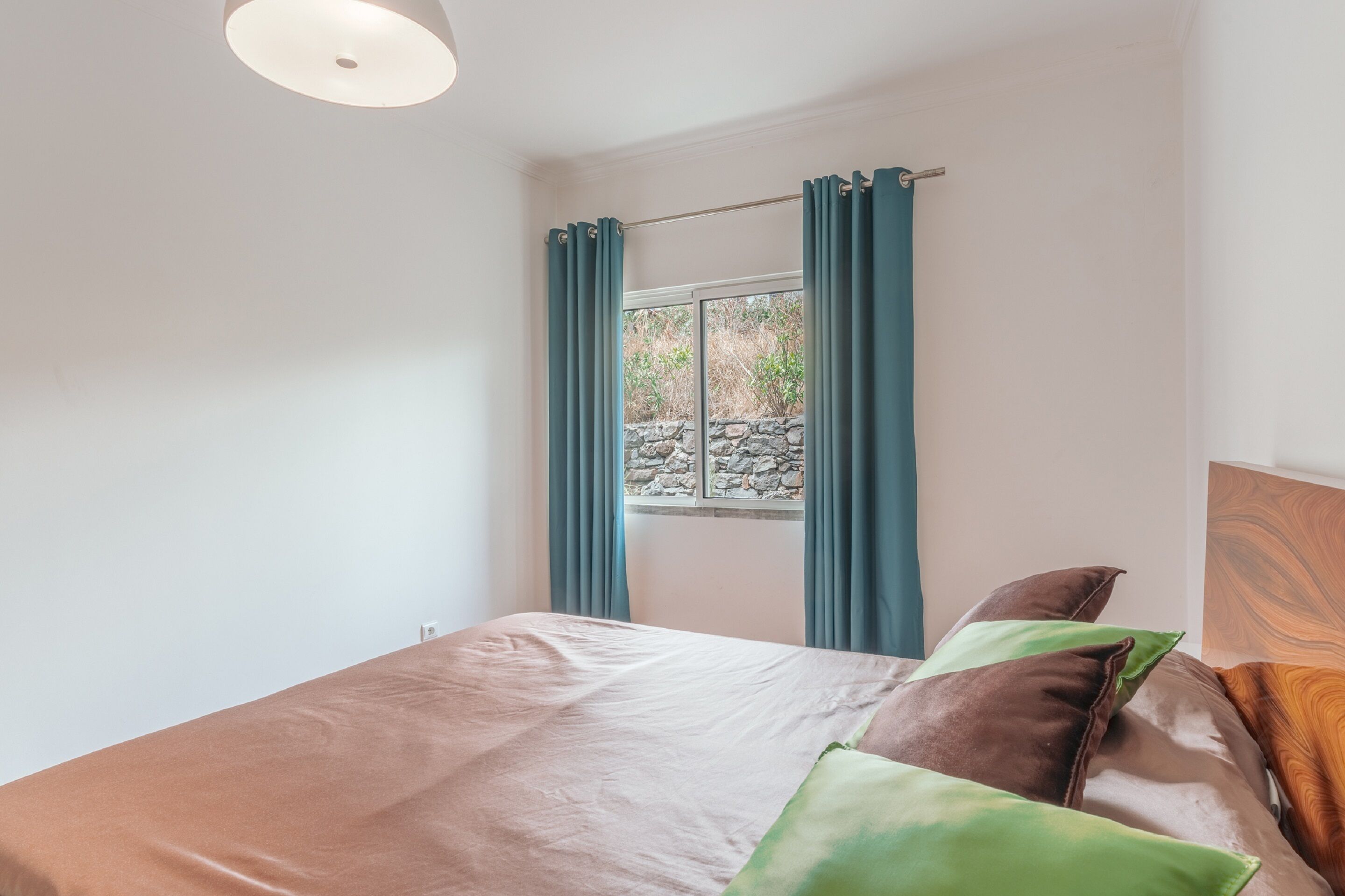Un dormitorio con cama, cortinas, ventana con vistas a una pared de piedra y cabecero de madera.