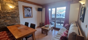 Un salon chaleureux avec un mur en pierre, une table à manger en bois, un canapé et un balcon offrant une vue sur des montagnes enneigées.