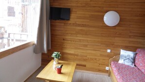 Une pièce aux murs recouverts de lambris en bois, un téléviseur à écran plat fixé au mur, un luminaire rond blanc, une petite table avec une tasse rouge et une plante, et un canapé agrémenté de coussins à motifs.