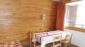 Una habitación con pared de madera, una cama con manta de cuadros, una mesa con mantel a rayas y una ventana con cortinas.