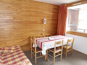 A room with a wooden wall, a bed with a checkered blanket, a table with a striped tablecloth, and a window with curtains. - travelski home classic - Résidence Saint Jacques - 2 pièces 5 personnes (La Plagne)