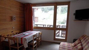 Una habitación con una mesa y sillas de madera, una cama con una colcha a cuadros, un televisor en la pared y una ventana con vistas a una terraza de madera y árboles.
