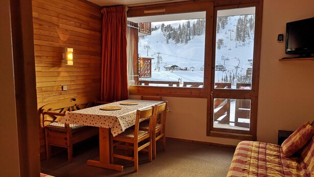 Una cabaña acogedora con interior de madera, una mesa de comedor con sillas, un sofá y un amplio ventanal con vistas a un paisaje montañoso nevado.