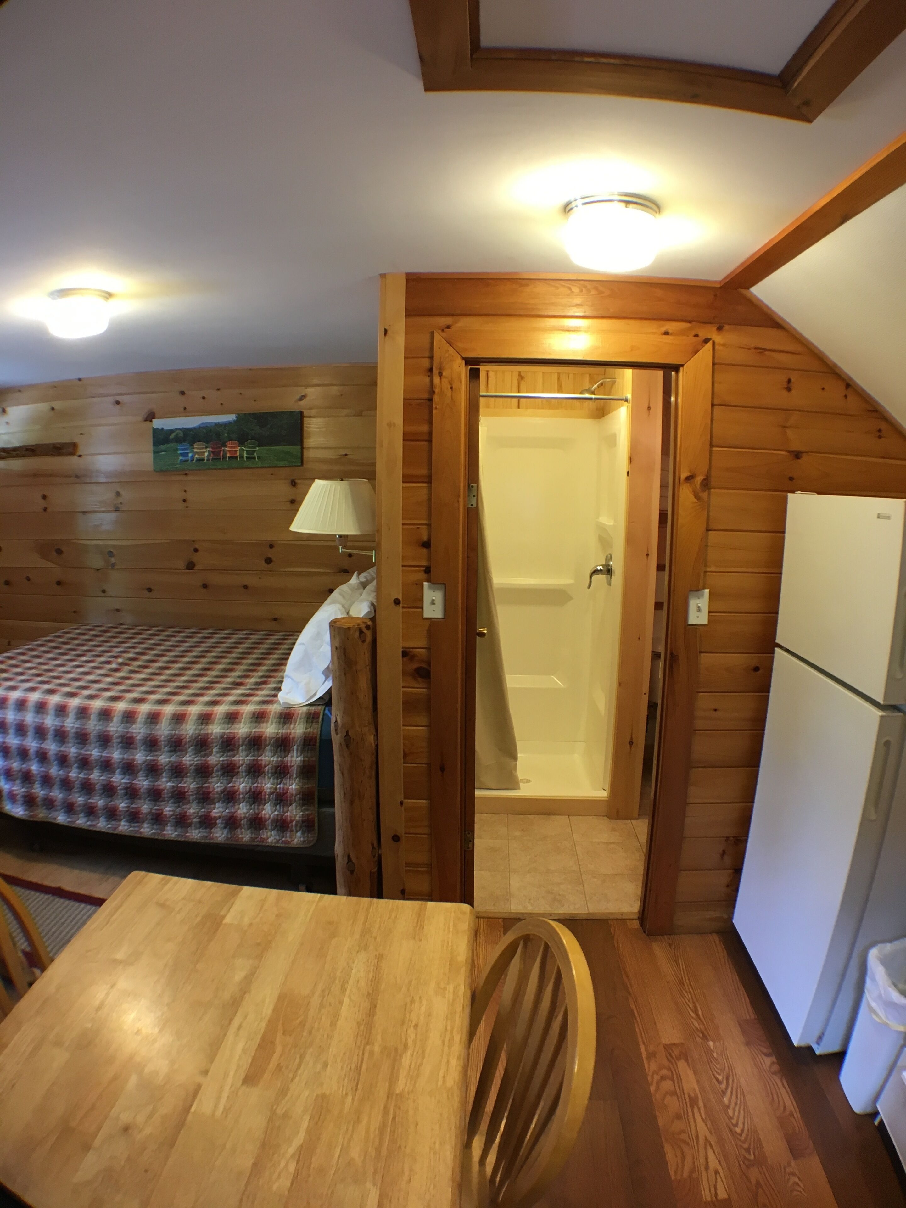 A wooden cabin interior with a bed, a table, a chair, a refrigerator, and a bathroom with a shower.