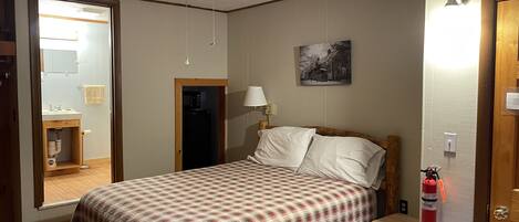 A bedroom with a bed, a ceiling fan, a nightstand, a lamp, a fire extinguisher, and a bathroom visible through an open door.