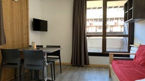 Una habitación con pared de madera, un televisor montado, una mesa de comedor con sillas, una ventana con cortinas y una cama con una colcha roja.