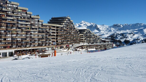 Un hotel de varios pisos con balcones, una pista de esquí en primer plano y montañas nevadas al fondo.