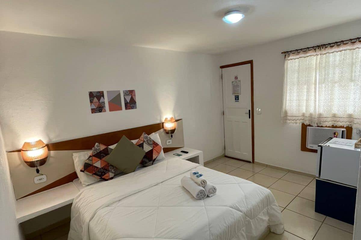 A hotel room with a bed, bedside lamps, a small table, a door, and a window with curtains.