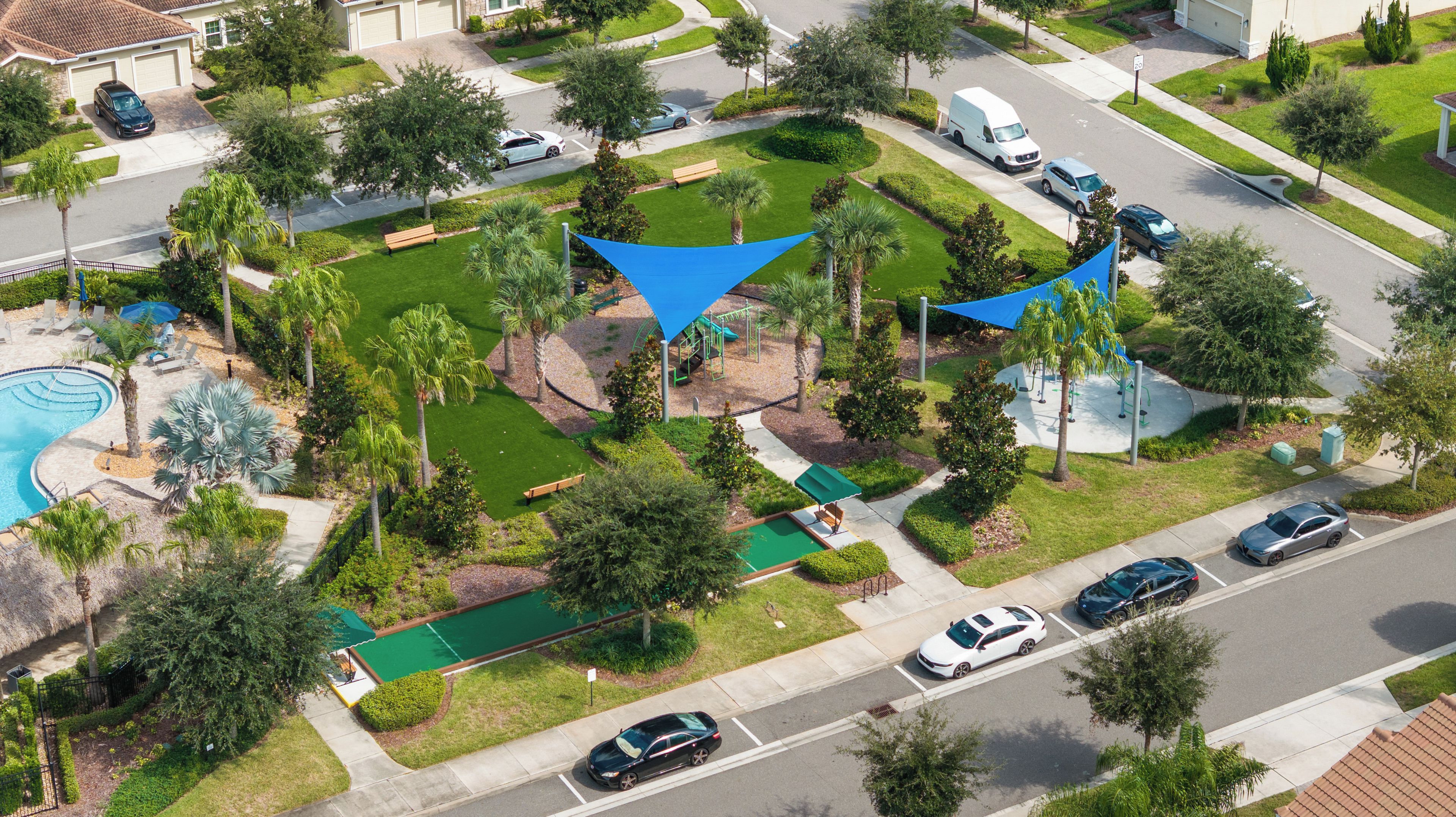 A residential area with a swimming pool, a playground, and a road with parked cars.