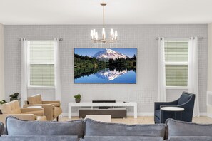 A modern living room with a flat-screen TV mounted on a white brick wall, a chandelier, and a large window with blinds.