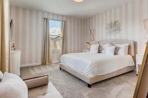 Ein Schlafzimmer mit einem großen Bett, einem Nachttisch, einer Lampe, einem Sessel und einem Fenster mit Vorhängen.