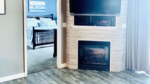 Una sala de estar moderna con un televisor de pantalla plana sobre una chimenea, un ventilador de techo y una puerta que da a un dormitorio.