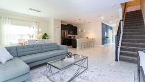 A modern living room with a grey sofa, a glass coffee table, and a kitchen in the background.