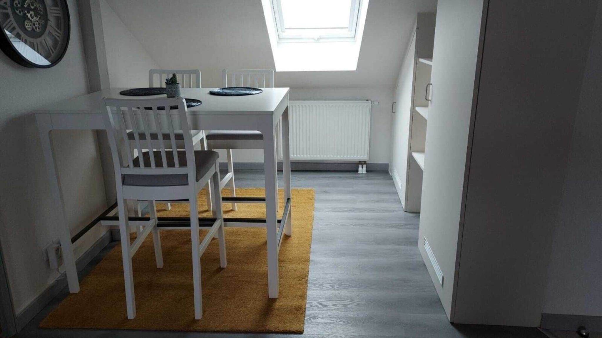 Una habitación compacta con una mesa y sillas blancas, una pequeña planta en maceta y un radiador.