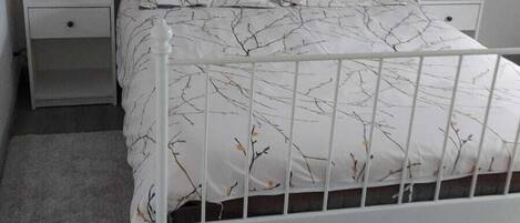 Un lit en métal blanc avec une tête de lit décorative, deux tables de chevet avec des lampes et un tapis gris sur un plancher en bois.