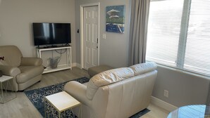 Un salon avec un canapé beige, une petite table d’appoint, un téléviseur à écran plat posé sur un meuble blanc, une porte et une fenêtre avec des stores.