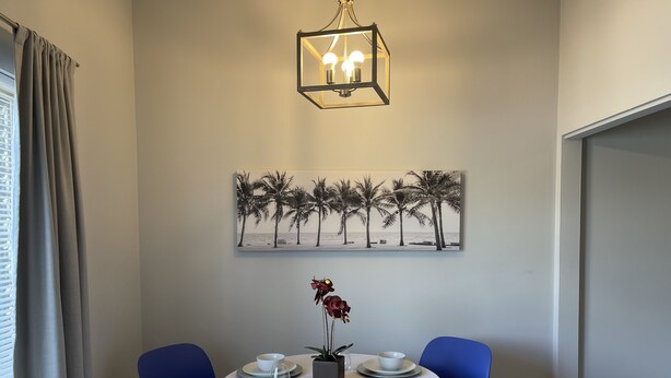 Un coin repas avec une table ronde, deux chaises bleues, une nappe blanche, une plante en pot et un tableau représentant des palmiers, accroché au mur.