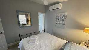 Un lit bien fait, avec une couette blanche et un oreiller gris, un ventilateur de plafond, un appareil de climatisation, un miroir encadré et un tableau accroché au mur.