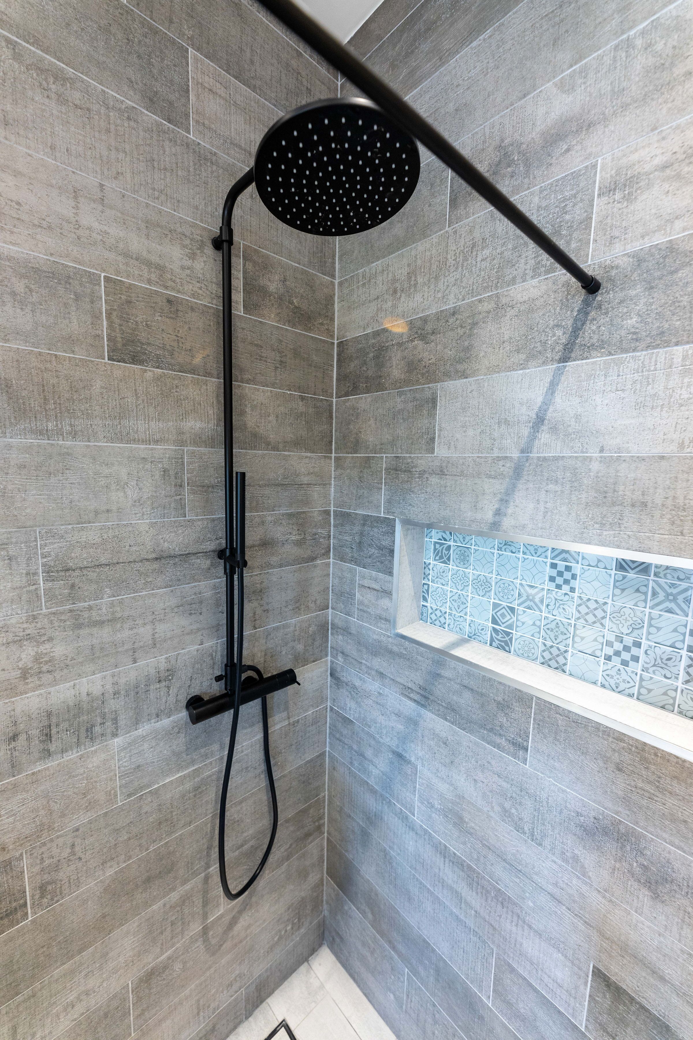 A shower with a black rain shower head and a handheld wand, set against a wall with grey tiles and a decorative tile shelf.