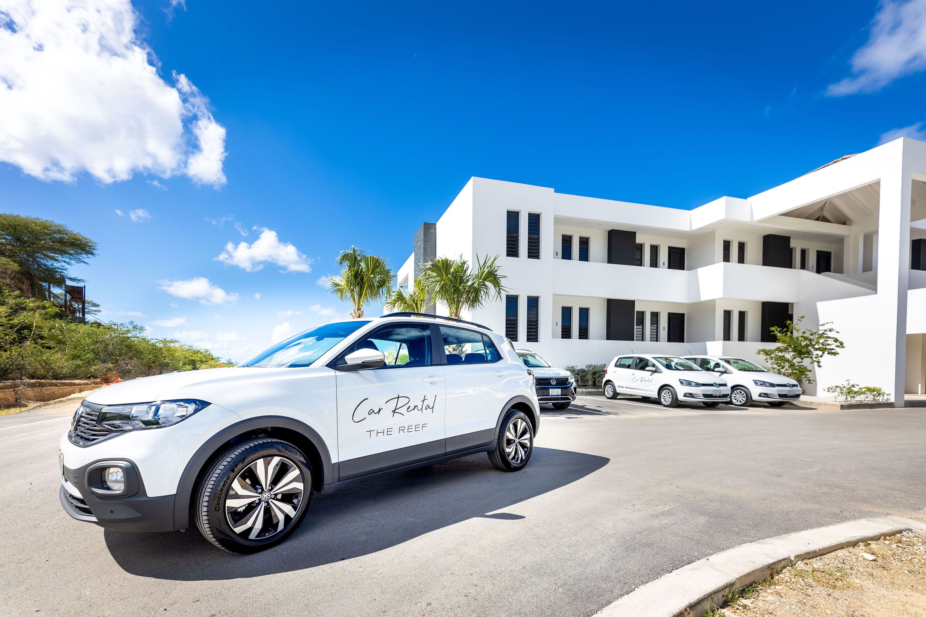 A white SUV with "Car Rental The Reef" on its side parked in front of a modern white building with multiple balconies.