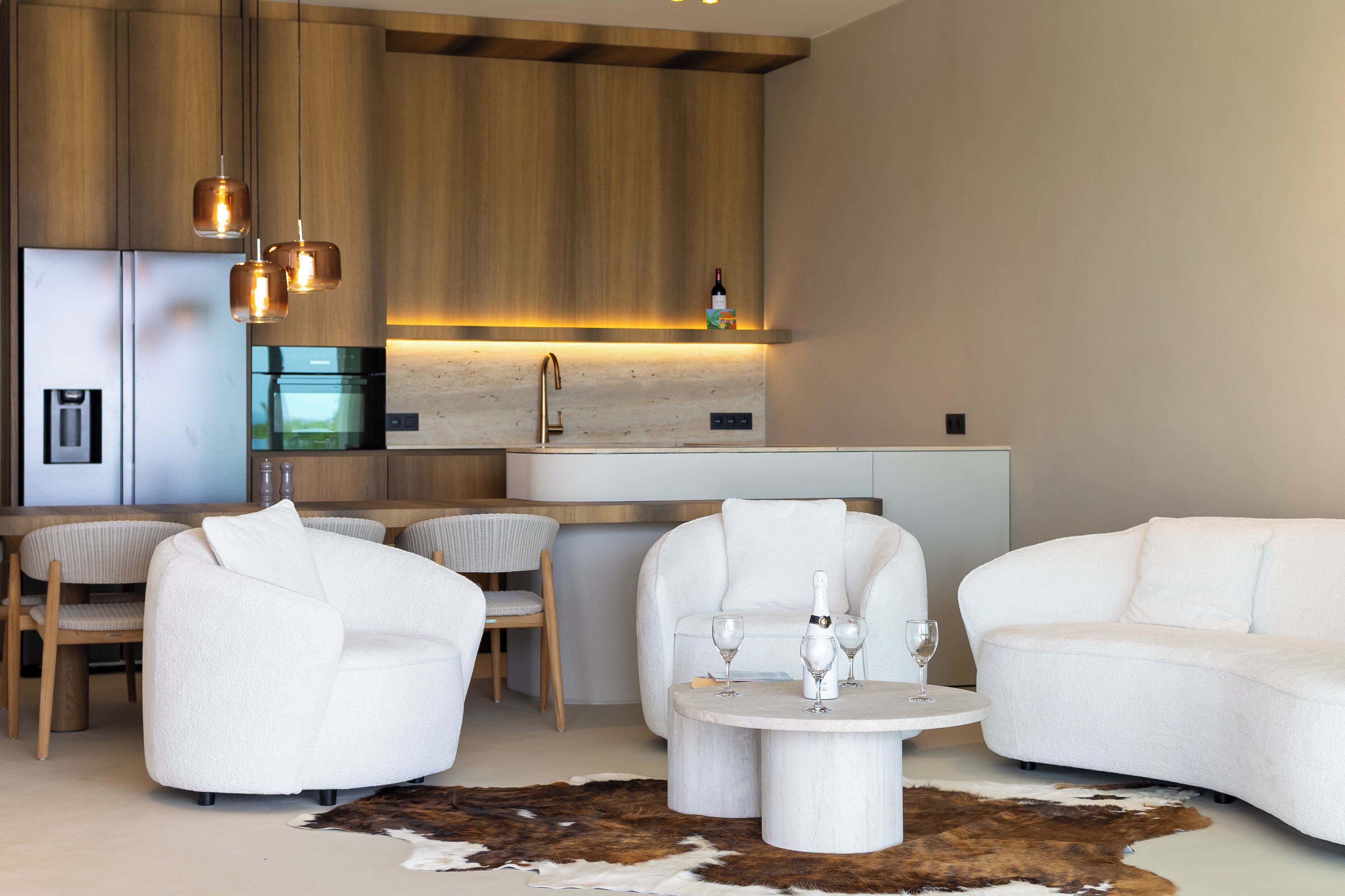 Eine moderne Küche mit Holzausstattung, einem Edelstahlkühlschrank und einem eingebauten Backofen. Zwei weiße Sofas, ein runder Couchtisch und ein Essbereich mit Stühlen sind ebenfalls vorhanden.