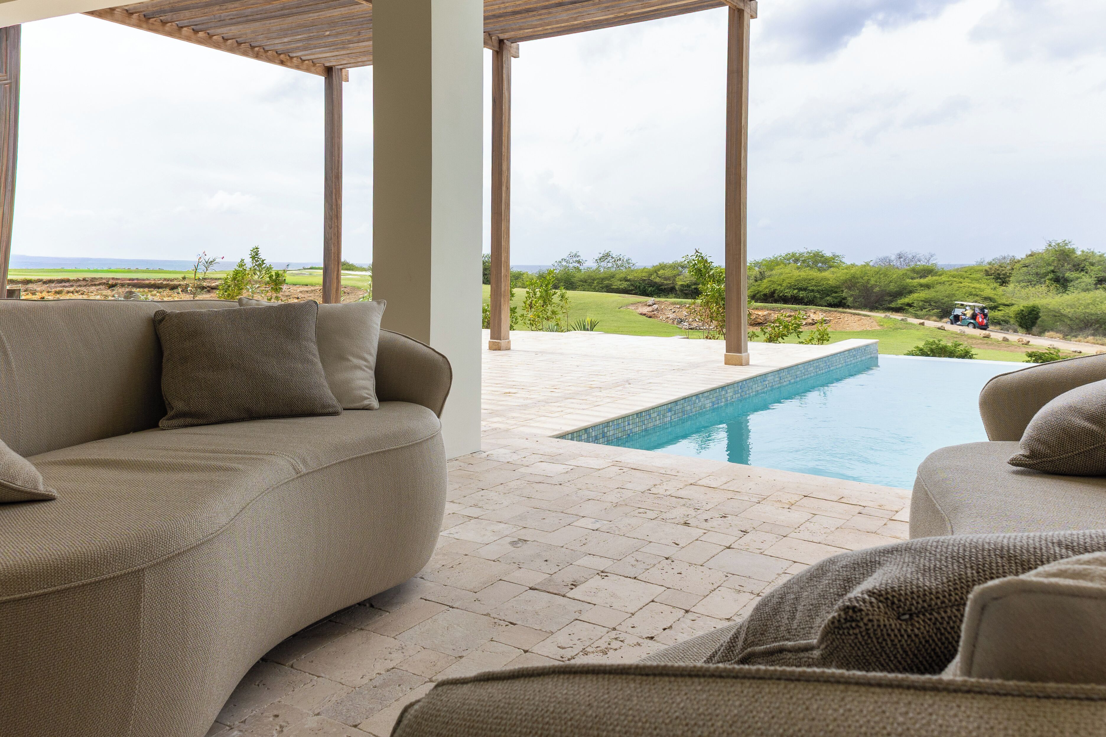 A covered outdoor area with a pool, beige sofas, and a view of a golf course.
