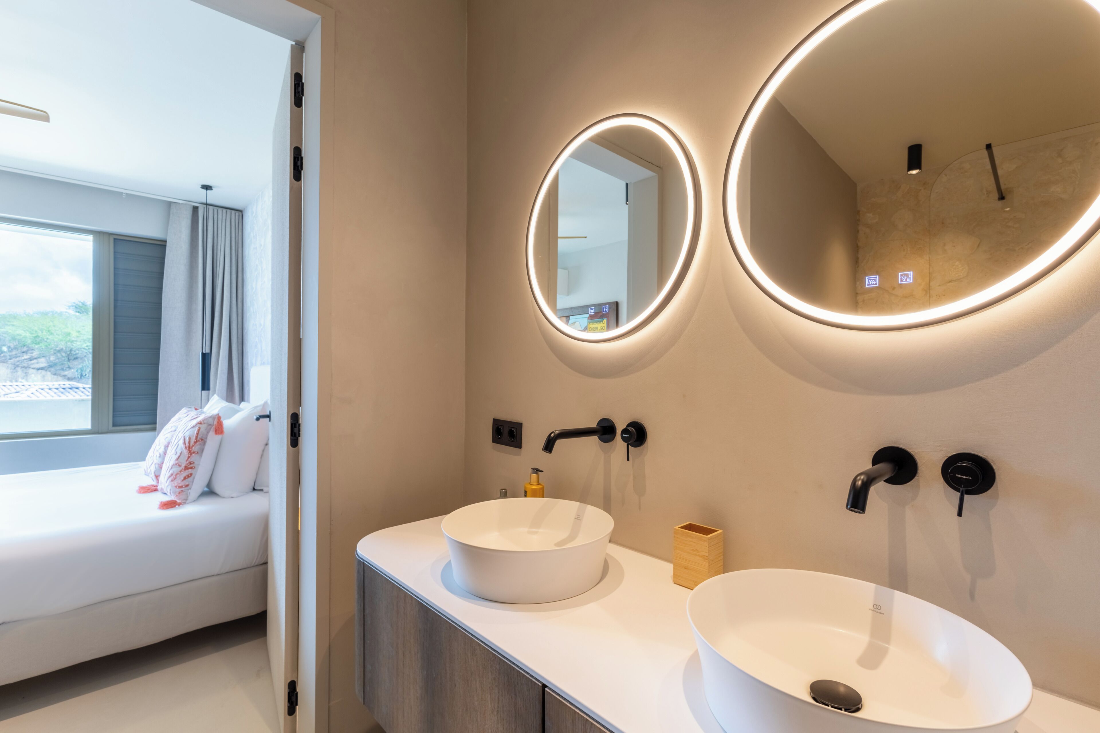 A modern bathroom with two round mirrors, a white sink, and a bedside table with a bed in the background.